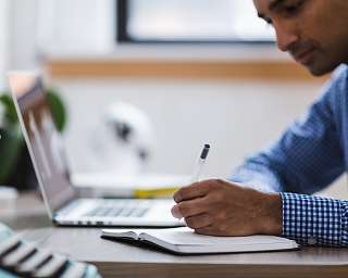 Online Lehrgang Person sitzt am Laptop und schreibt auf einem beiliegenden Schreibblock